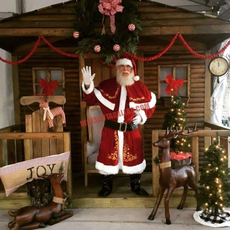 Real bearded Santa for hire waves from a wooden cabin set with wreaths, small trees, and reindeer figures in Dallas.