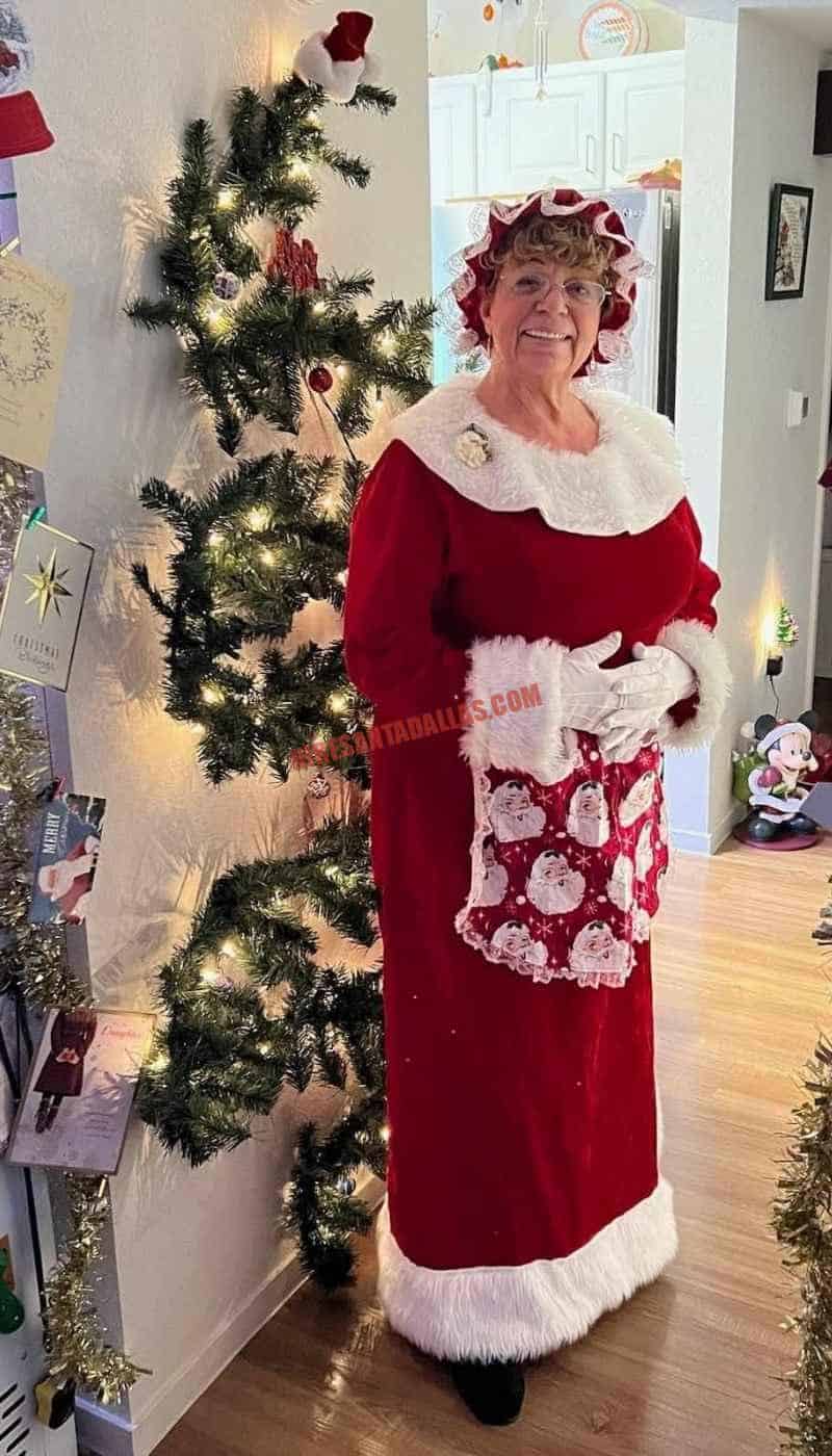 Mrs. Claus for hire in Dallas in a red dress, white fur cuffs, and a Santa-print apron beside a wall Christmas tree.