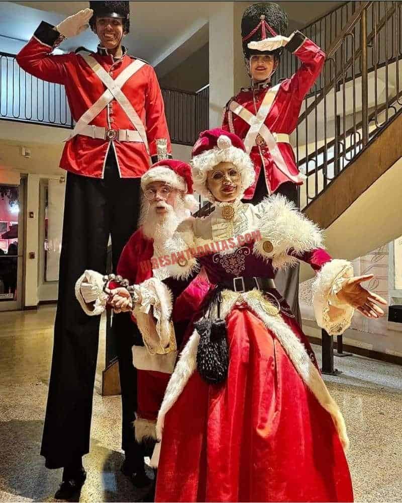 Corporate Santa in a lobby with Mrs. Claus and two nutcracker soldiers on stilts beside a staircase.