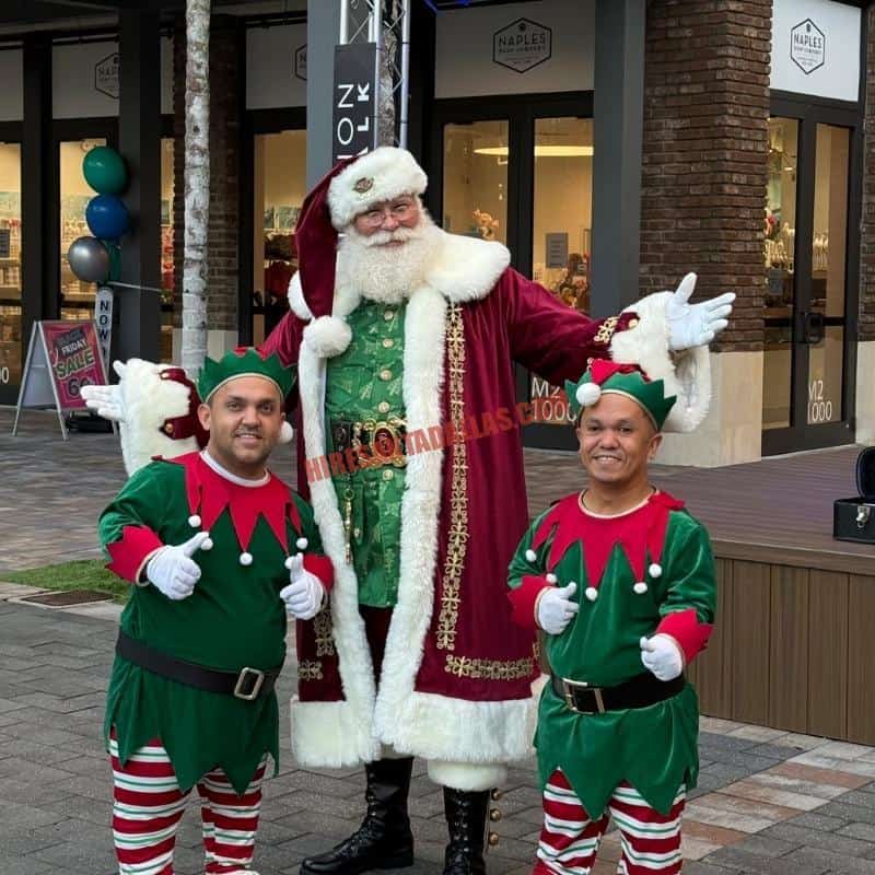Santa impersonator for hire in a burgundy coat and green vest stands with two elves at a brick plaza.