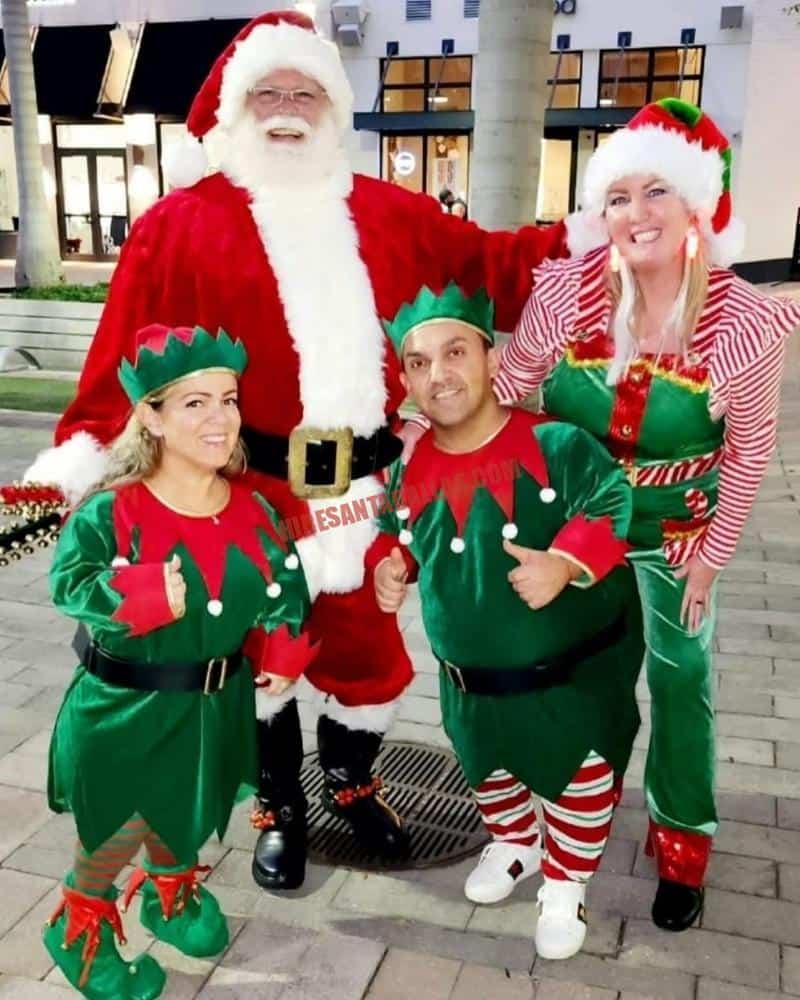 Hire Santa Irving: Santa in a red suit stands with three elves on a paved walk outside a building.