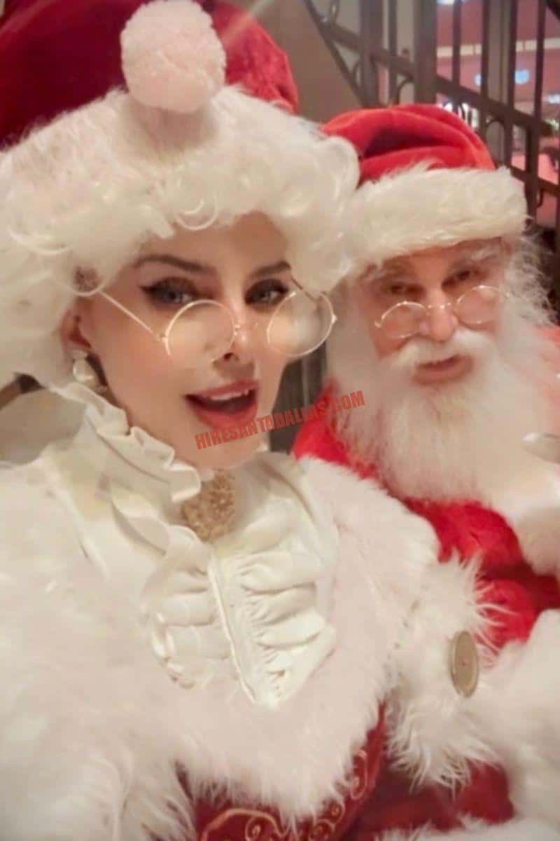 Santa and Mrs. Claus in red and white costumes pose together for a Dallas booking, she in ruffled white and he in a red suit.