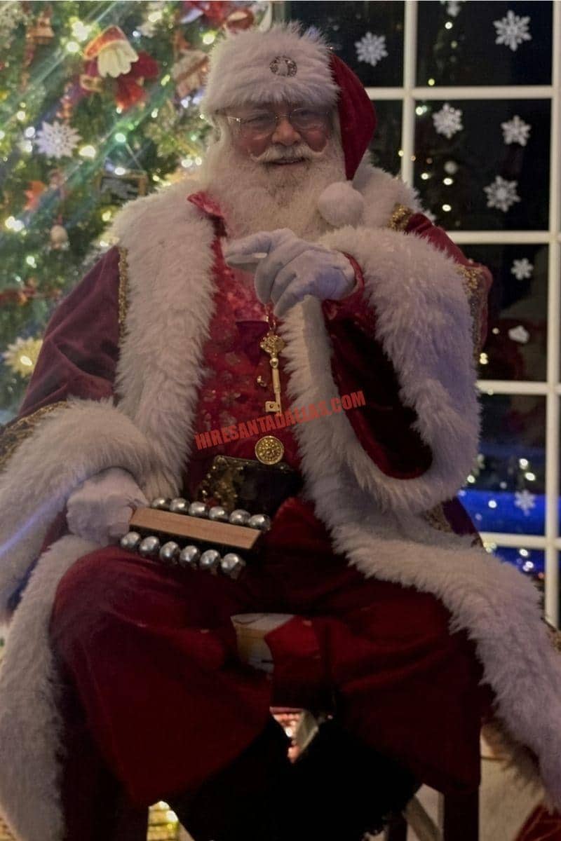 Santa home visit in Dallas: Santa in a burgundy coat holds sleigh bells beside a lit tree and a night window.
